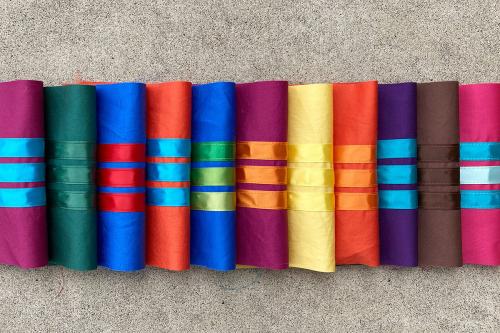 A stack of pieces of cloth, each a different vibrant color, each sewn with three ribbons across in contrasting colors.