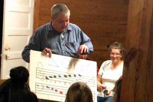 A man holds up a music chart that has simple geometric shapes in place of notes: triangle, circle, square, diamond. 