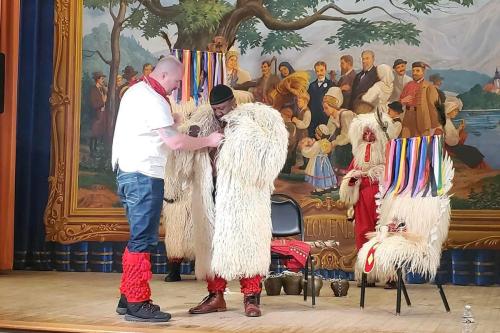 On an indoor stage, one man helps another suit up in a furry sheepskin.