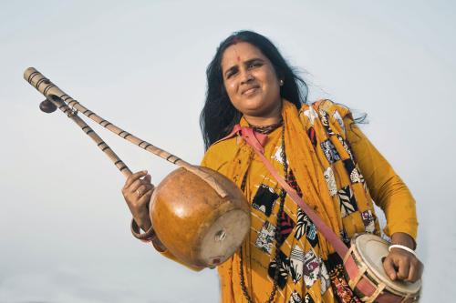 A woman in a yellow tunic holds up a wooden stringed instrument in one hand and a small drum in the other.