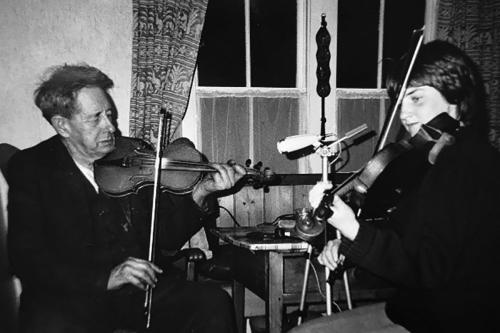Two people play fiddles, sitting across from each other inside a home.