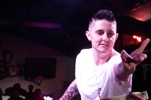 A person in a white T-shirt and a necklace bearing a cross, lit by stage lights, reaches out toward the camera. In the foreground, someone is holding a dollar bill out toward them.
