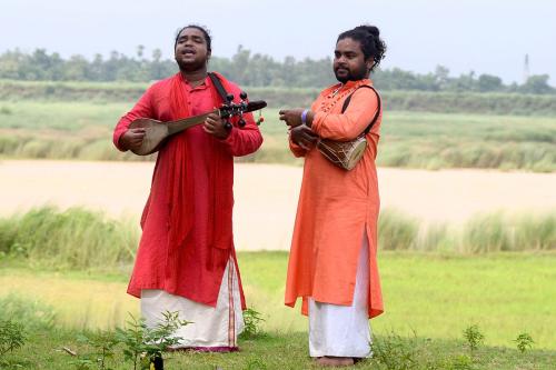 Two men, who appear to be twin brothers, play music with a vast green meadow behind them. The one of the left is in a red tunic, playing a wooden stringed instrument. The one of the right is wearing a bright orange tunic, playing a wooden drum tucked unde
