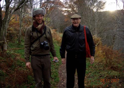 Two men stop to pose along a dirt hiking trail in the woods. One carries a camera. Date stamp in orange reads 06-11-2007.