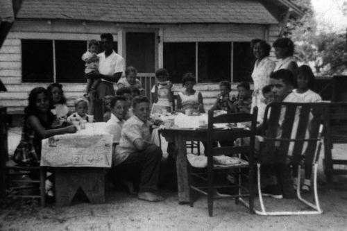 A group of people at tables in a backyard. Black-and-white photograph.