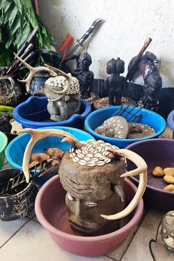 Various objects arranged in purple and blue bowls on the ground. Two hold stone carved like human heads but with shells for eyes and hair and antlers. Others hold small black figurines. Others hold food.
