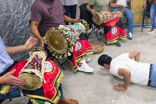 In front of three seated drummers, their heads out of frame, a man lays face-down on the ground, resting on his palms.