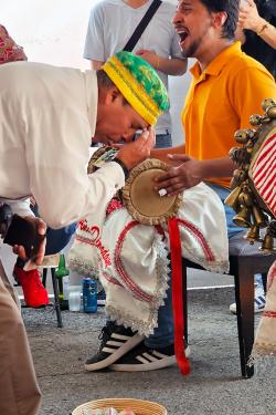 One man bows over a woven basket, holding his fingers to his forehead, with eyes closed. Behind him, another man plays a double-headed drum in his hap, mouth wide as he sings.