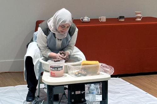 A woman leans over a potter wheel, sculpting a piece of reddish-brown clay.