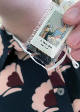 Closeup on a hand holding up a small rectangular picture, labeled St. Simon Stock, in a plastic slip and hanging on a white cord around her neck.