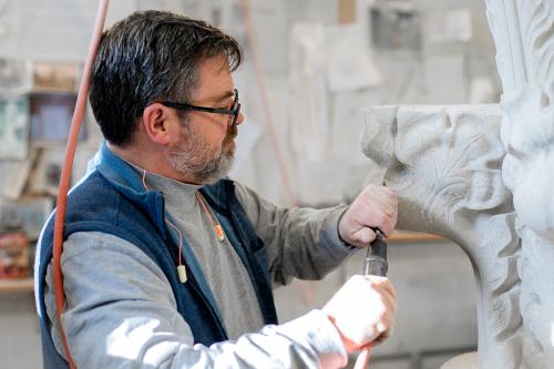 A man faces a white stone sculpture, holding a pneumatic chisel to an ornamental design in the surface. 