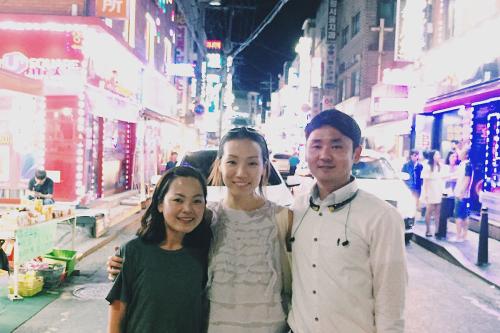 Three people pose, arms around each other, in the middle of a street lit by neon storefront signs.
