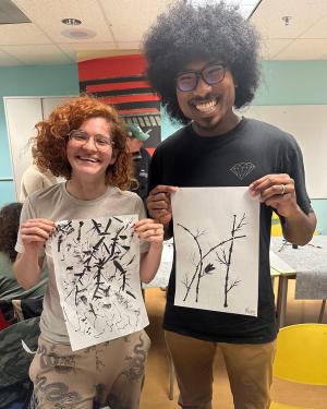 Two people, one with peach-colored skin and curly red hair and one with dark skin and black curly hair, smile widely as they each hold up a sheet of paper with paintings of bamboo plants.