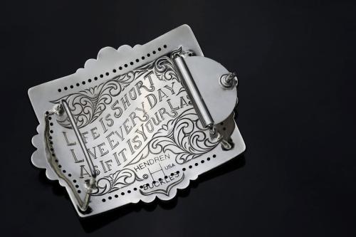Closeup on the back of a silver belt buckle, engraved with the words: Life is short. Live every day as if it is your last. Copyright Hendren Buckles, USA.