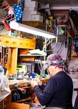 A man works in a cluttered workshop under a bright light, wearing a magnifying headset.