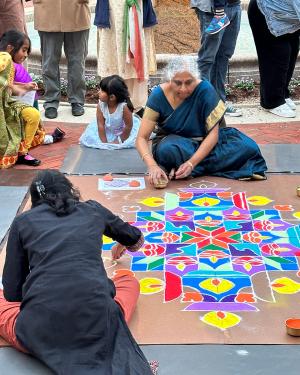 The two women, sitting on the ground, continue their mandala design, radiating out from a central orange and red flower shape. In reds, blues, and purples, there is a repeating design of a lit candle in a shallow bowl. Kids sit around the artists.