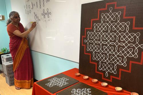 A woman in red and orange wrap dress draws a curving line on a whiteboard. On display next to her are completed patterns with this curving lines, punctuated by dots, painted in white and bordered in red on a black background.