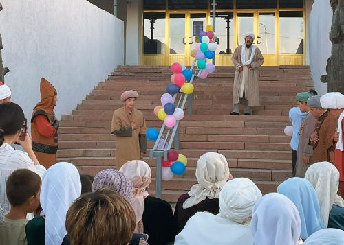 A crowd of people stand on and around the stairway entry to a building, with their hands over their hearts. The central hand rail is decorated with clusters of colorful balloons.