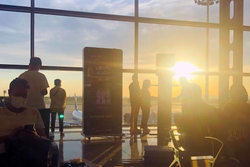 The sun rising or setting outside the window of an airport terminal, with a few people waiting.