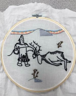 Embroidery on white cloth, still in the hoop, depicting a scene of a person hunting a caribou.