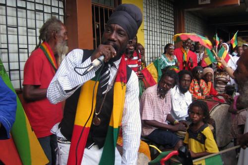 A man singing into a microphone in front of a crowd of people sitting and standing, most wearing or holding flags in the colors reg, yellow, and green.