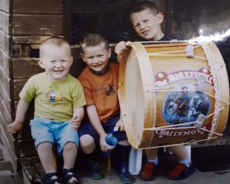 Three young boys sit and smile, one holding a large cylindrical drum. Old color family photo.