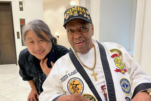 A woman and a man, who is dressed in a white gi garment with various patches, a gold cross necklace, and a black hat embroidered with the word SIMBA and adorned with several buttons, pose smiling.