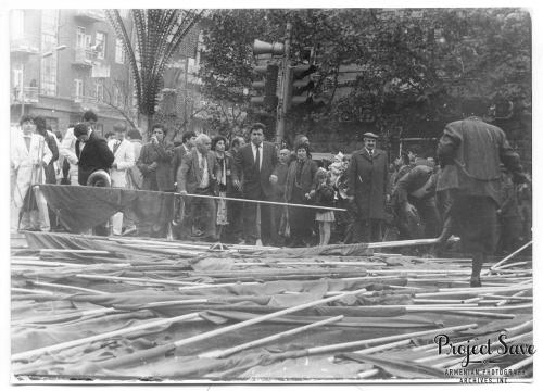 In the foreground, dozens of flag poles laid on the ground. One person navigates over them, toward a crowd of people in the background. Black-and-white photo, with a watermark for Project Save in the lower right corner.