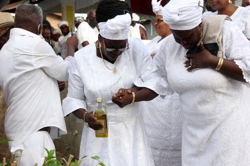 Two women, both in white dresses and headwraps, clutch hands and face downward in prayer. One holds an open bottle of olive oil. Behind them, a crowd also dressed in white.
