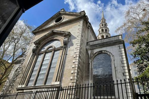 Exterior of a church made of gray bricks, with a black wrought-iron fence in front and a pointed spire rising in the background.