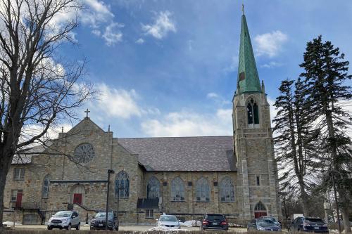 Exterior of a gray and brown stone church with a tall green steeple and cars parked in front.