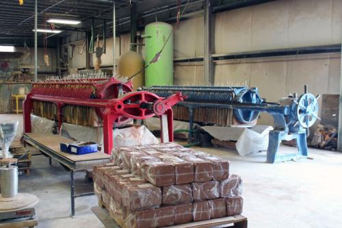 Interior of a factory workroom with blocks of plastic-wrapped clay on a pallet in the foreground and large machinery in the background.