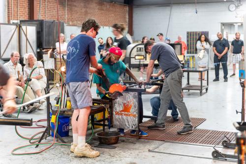 Four people work together to form a blob of red-hot glass while a crowd around them watches.
