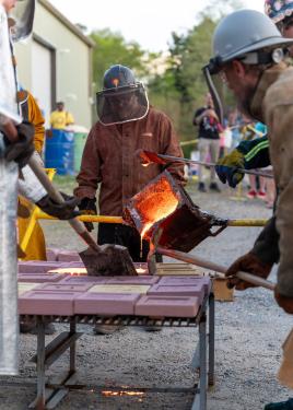 Four people handle a container of molten metal, pouring it among bricks on a grate outdoors.