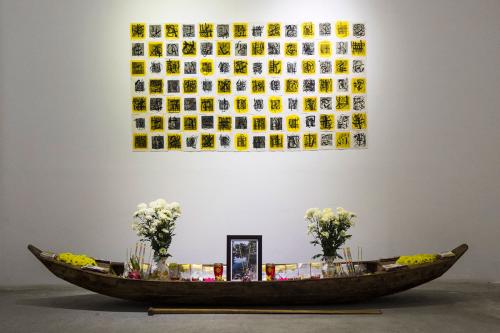 An art installation in a gallery: on the ground, a canoe-shaped vessel holdings bouquets of white and yellow flowers, a framed photo, incense sticks, and other items. Above it, on the wall, is a grid of alternating white and yellow squares, each with a bl