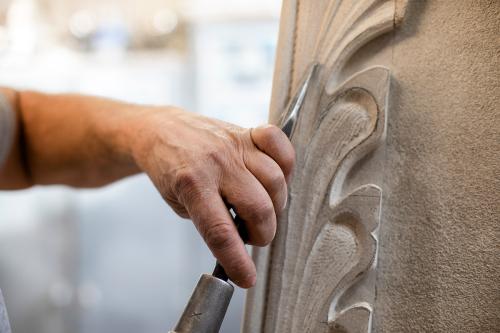 Close-up of a hand holding a chisel, pointed upwards and ready to carve intoa piece of white, ornate stone on the right.