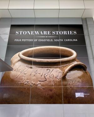 Large poster with an image of a ceramic jug, with an inscription in the top, including the name Dave. Text on the poster reads: STONEWARE STORIES: FOLK POTTERY OF EDGEFIELD, SOUTH CAROLINA. SFO Museum logo in the bottom right corner.