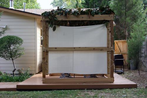 Small outdoor structure made of wooden beams, white fabric walls, and a roof thatched with dense greenery. It appears to be on a patio in the backyard of a house.