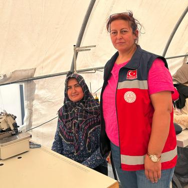 Two women pose, one with a headscrarf sitting at a sewing machine, and the other in a red vest standing beside her.