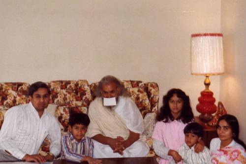 Old family photo with the Jain family and an older man posing, sitting on and around a beige floral-patterned couch. The older man is wearing a white face mask.