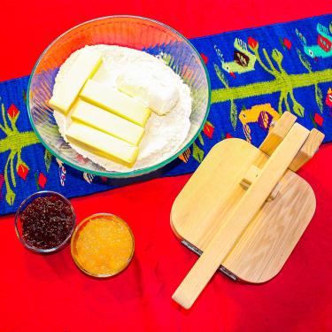 From above, bowls of dark red and orange jam, a bowl with flour, four sticks of butter, and a block of cream cheese, and a wooden tortilla press.