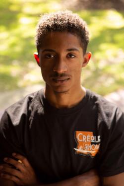 A young Black man poses with his arms crossed. His T-shirt has a logo for Créole Show, with the shape of the state of Louisiana.