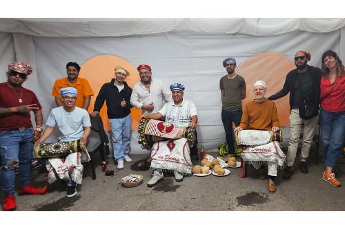 Nine men and one woman pose under the tent, three holding drums in their laps. One gives a thumbs up, and another a peace sign.