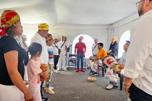 UNder a white tent with arched “windows,” a group of men and women circle around, singing and playing drums.
