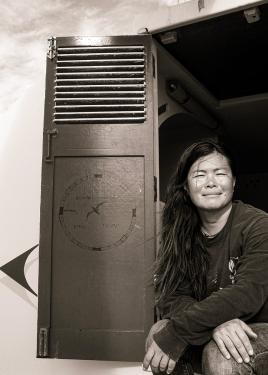 A woman with long dark hair sits in a doorway of a ship, squinting toward the camera. Black-and-white photo. Painted on the open door next to her is the Hawaiian compass rose.