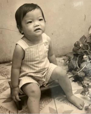 A baby with dark hair and eyes, wearing a striped jumper, sits on the ground, looking up. Black-and-white photo.