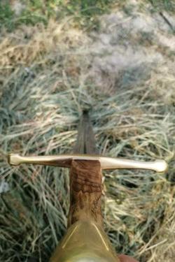 Close-up on the bronze-colored handle of a sword, the edge of which sticks into long grass. 