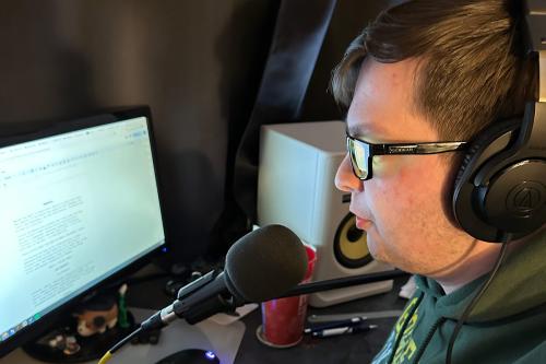 A man in a green hoodie and headphones looks at a computer screen, a microphone positioned in front of his mouth.