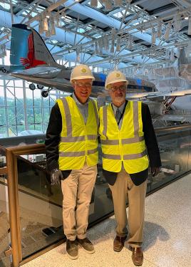 Two men wearing yellow safety vests and white hard hats with the Smithsonian logo pose in front of an airplane suspended from the ceiling in a museum.