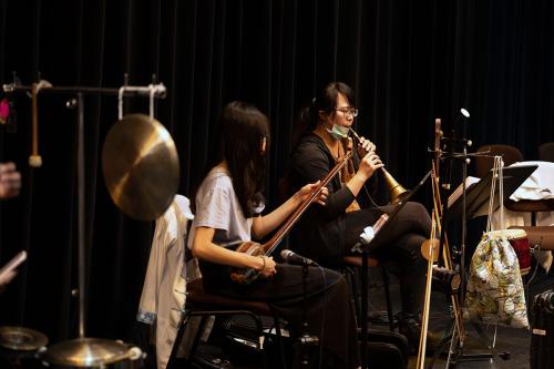 Two members of a small orchestra play backstage, one on a long-necked lute and another on a double-reed wind instrument. A small gong hangs in the foreground.